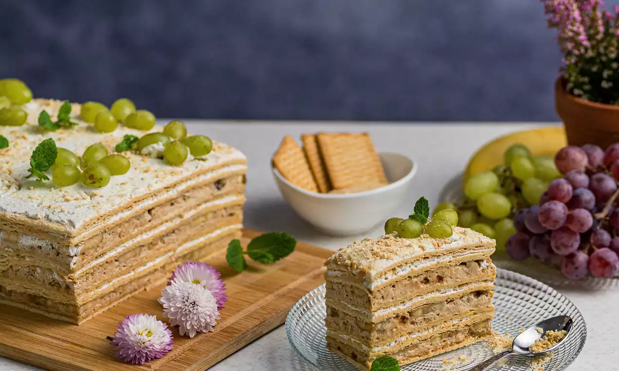 Prăjitură stratificată cu cremă fină și biscuiți aurii, decorată cu struguri verzi, mentă proaspătă și flori pe un platou elegant