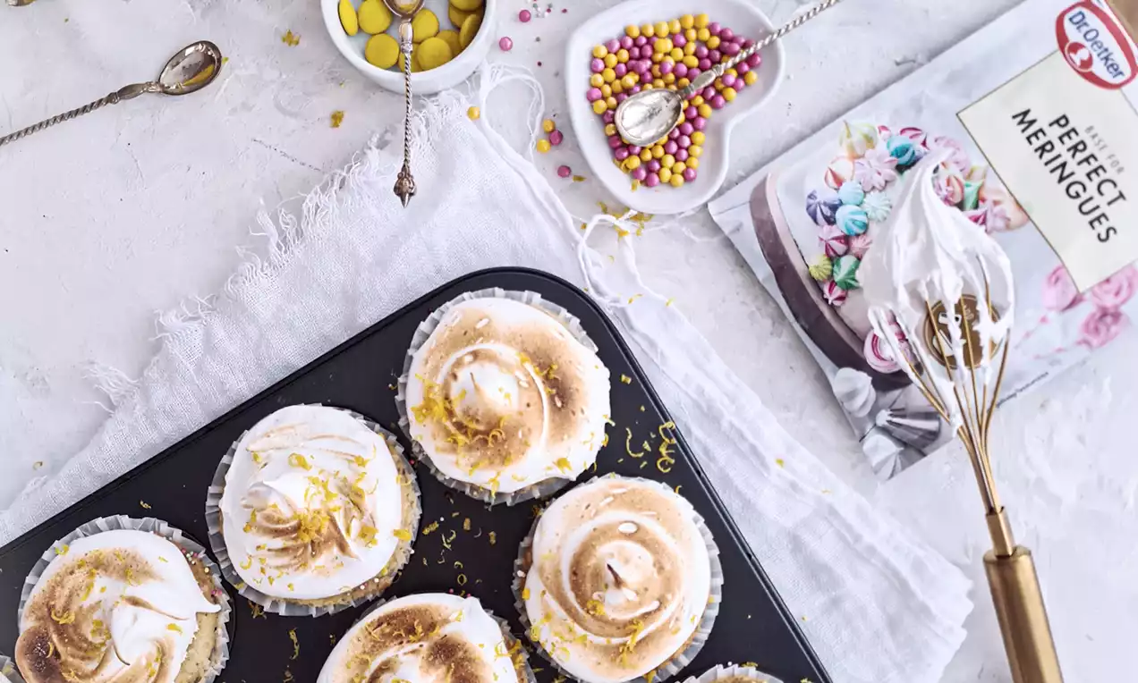 Citronmuffins med gyllene marängtoppar, rivet citronskal och färgglada strössel bredvid skål med citroner och dekorationer på ljus bakgrund