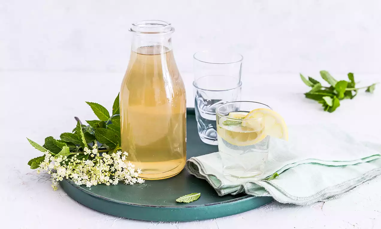 Světle zlatavý sirup v průhledné lahvi s jemným leskem, doplněný sklenicí vody s plátkem citronu a lístkem máty na zeleném podnose