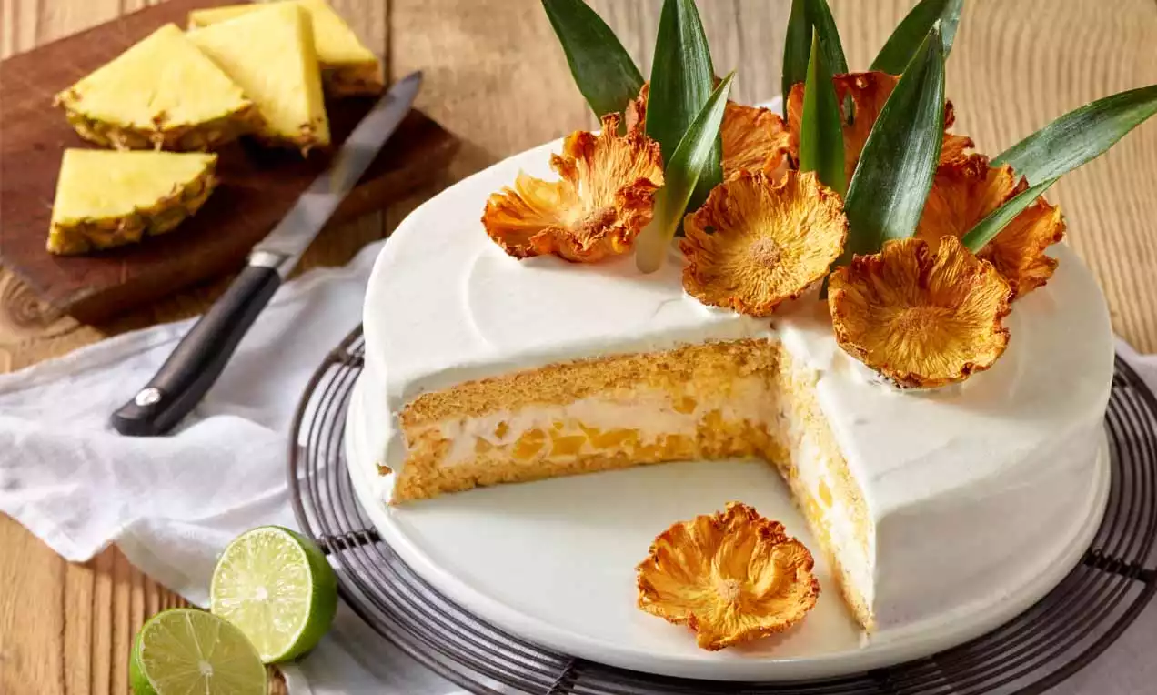 Joghurt-Ananas-Torte mit weißer Creme, gelben Ananasstücken und knusprigen Ananasscheiben, garniert mit grünen Blättern auf einer Tortenplatte