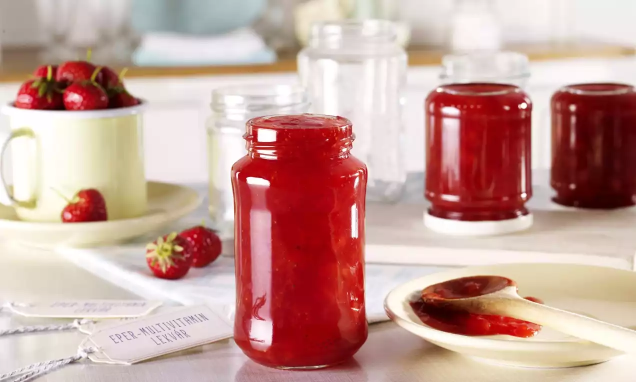 Egy befőttesüveg élénkpiros epermultivitamin lekvár sima textúrával, mellette fa kanál, tányér, friss eprek és további üvegek a háttérben