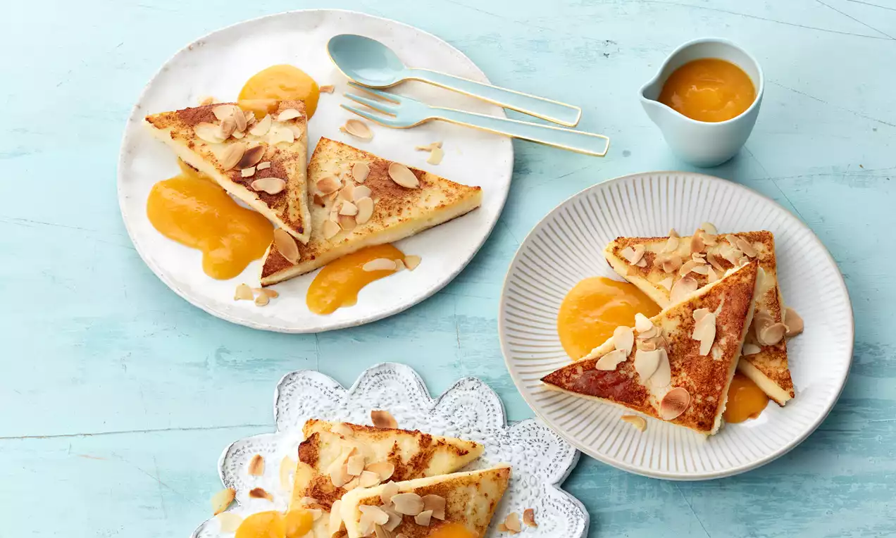 Goldbraune Grießschnitten mit leichter Bräunung, mit Mandelsplittern bestreut, dazu leuchtend orange Fruchtpüree auf weißem Teller