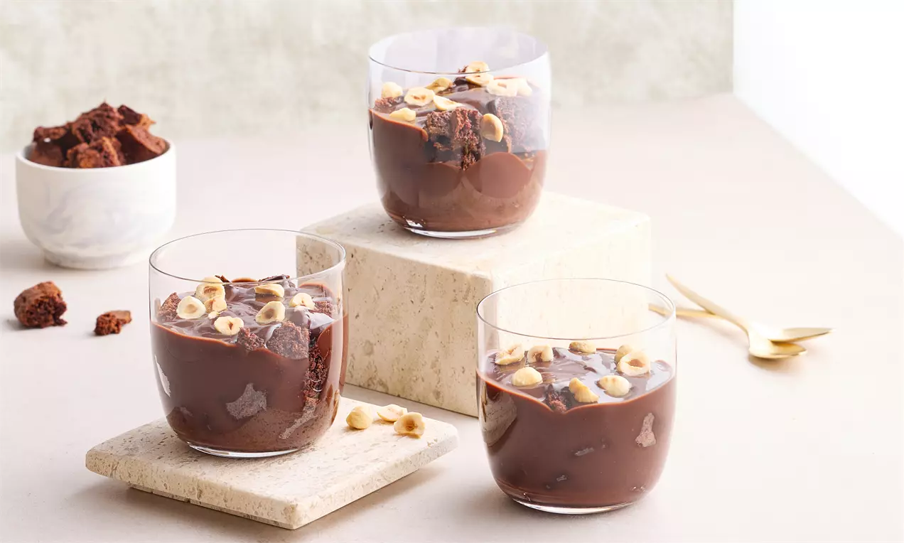 Cam bardaklarda sunulan yoğun çikolatalı puding, kek parçaları ve kavrulmuş fındıklarla dekore edilmiş, doğal tonlarda bir sunum