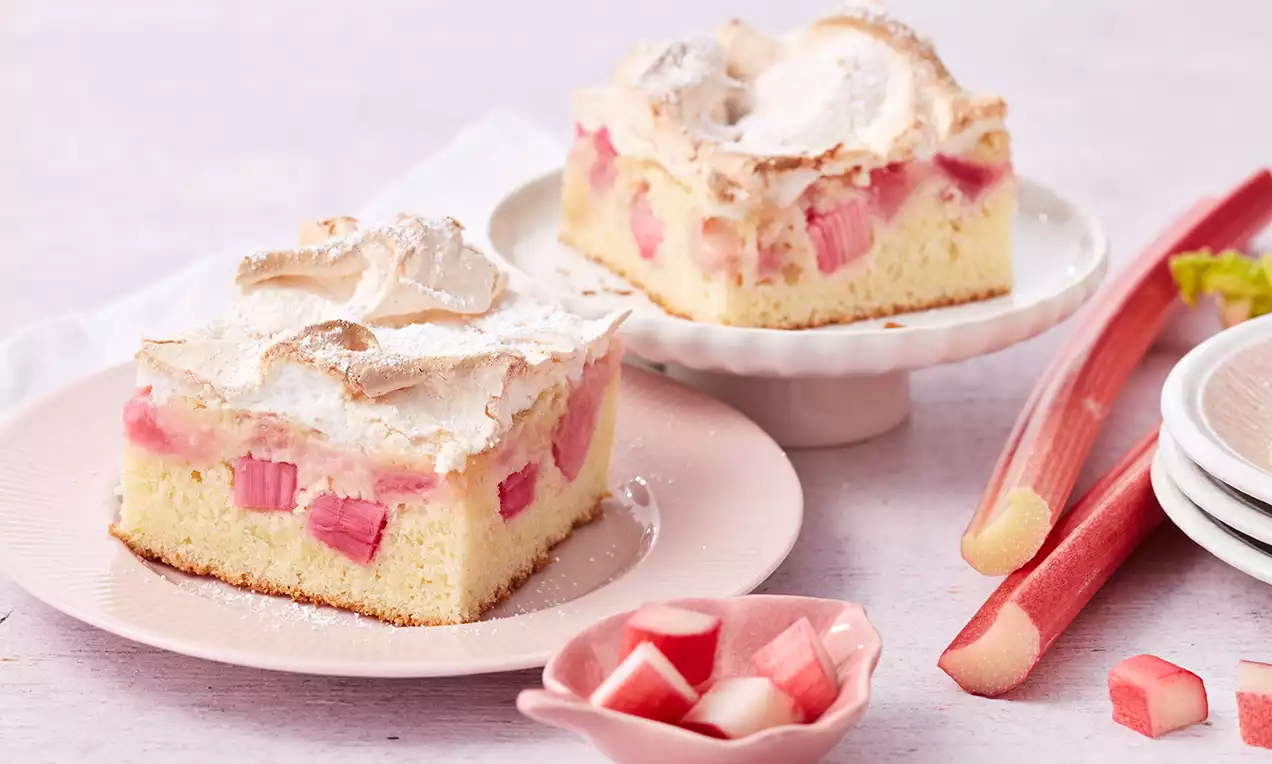 Zwei Stücke Rhabarberkuchen mit goldbrauner Baiserhaube und rosa Rhabarberstücken auf Tellern, dekoriert mit frischem Rhabarber