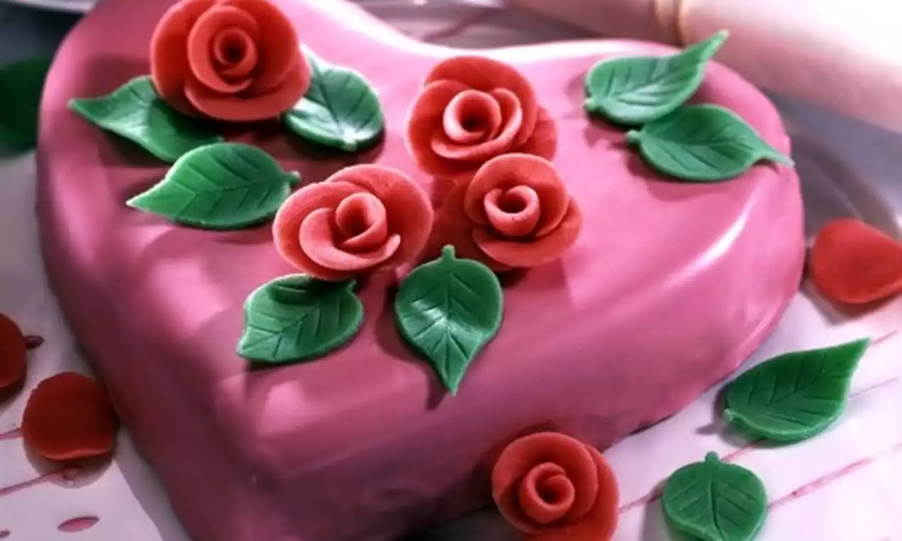 Gâteau en forme de cœur rose brillant décoré de roses rouges en pâte avec des feuilles vertes sur une assiette blanche
