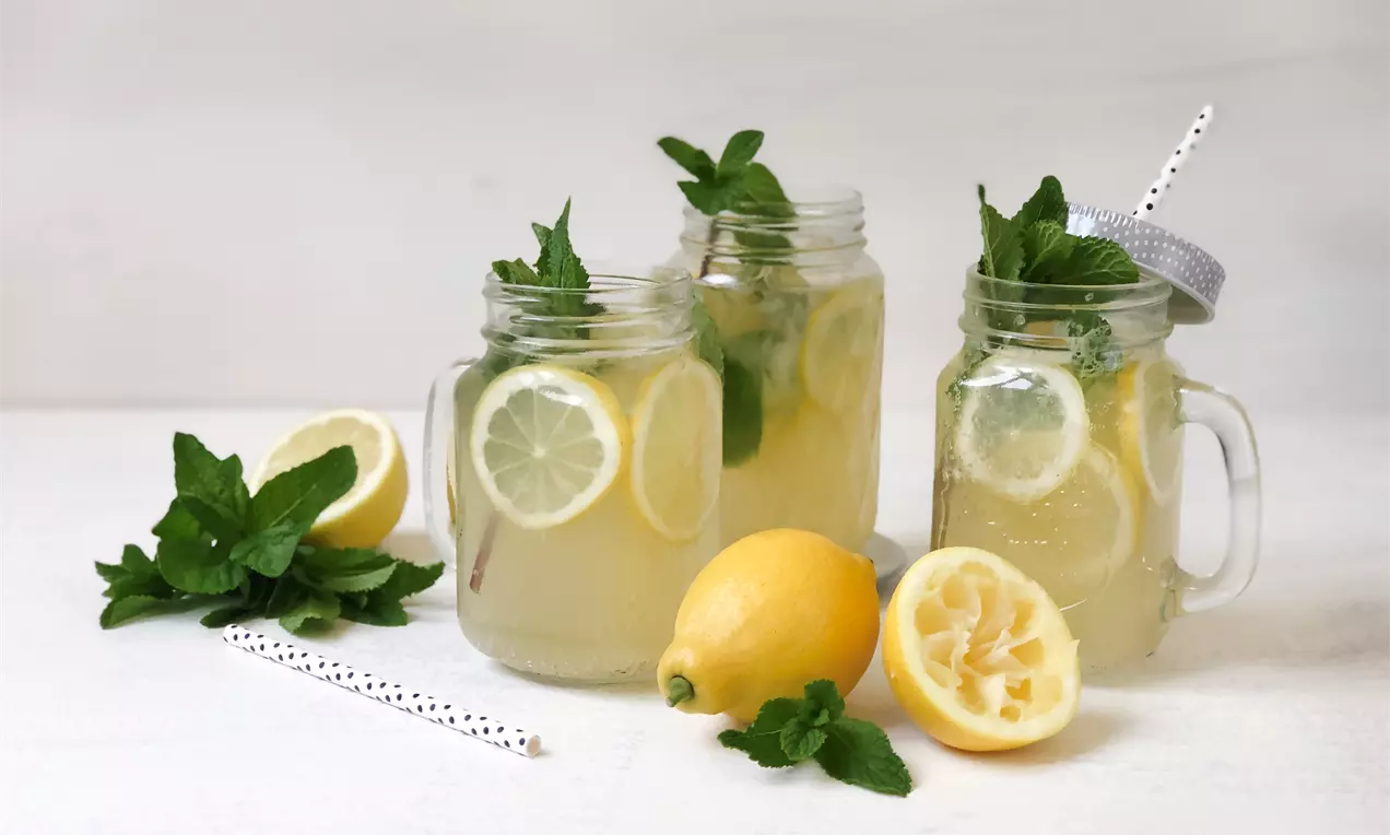 Citrónovo-mätová limonáda v priesvitných pohároch s uškami, ozdobená plátkami citróna, čerstvou mätou a papierovými slamkami na bielom podklade