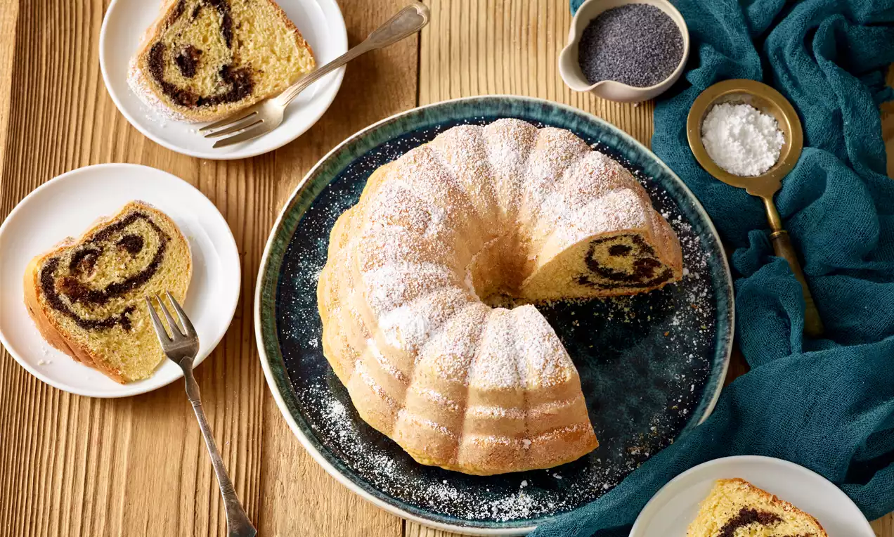 Gerollter Gugelhupf aus hellem Teig mit dunkler Mohnfüllung, leicht mit Puderzucker bestäubt auf blauem Teller, umgeben von kleineren Tellern