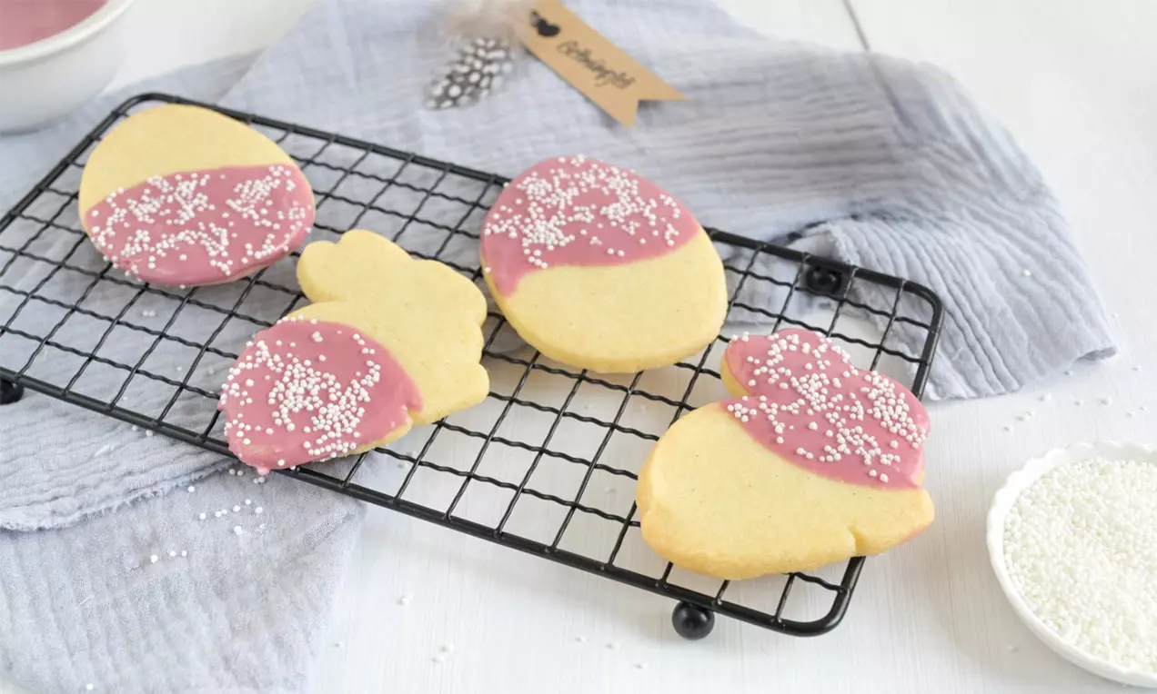 Buttergelbe Shortbread-Kekse mit halbseitiger rosa Glasur und weißen Zuckerstreuseln, arrangiert auf einem Metallgitter mit Stoffunterlage