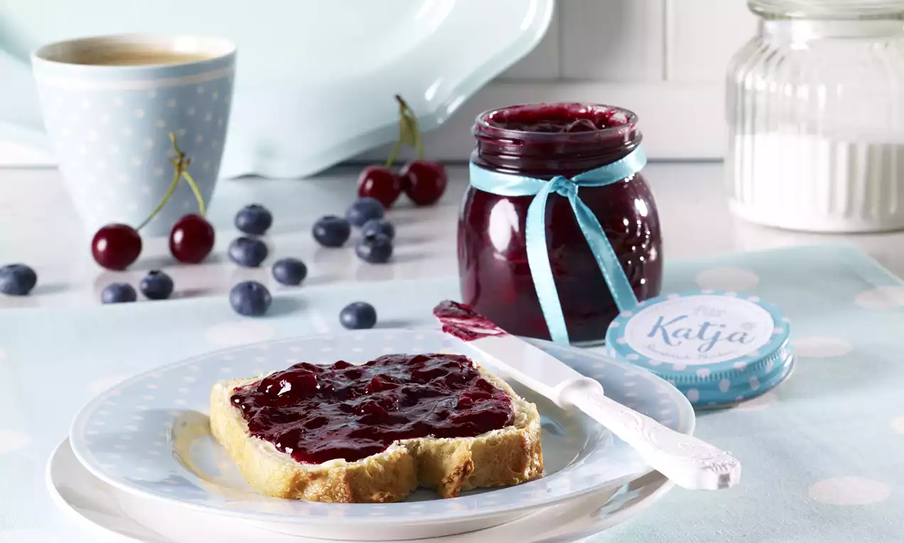 Ein Glas mit tiefroter Konfitüre, verziert mit blauem Schleifenband, daneben ein mit Konfitüre bestrichener Brotscheiben auf Teller
