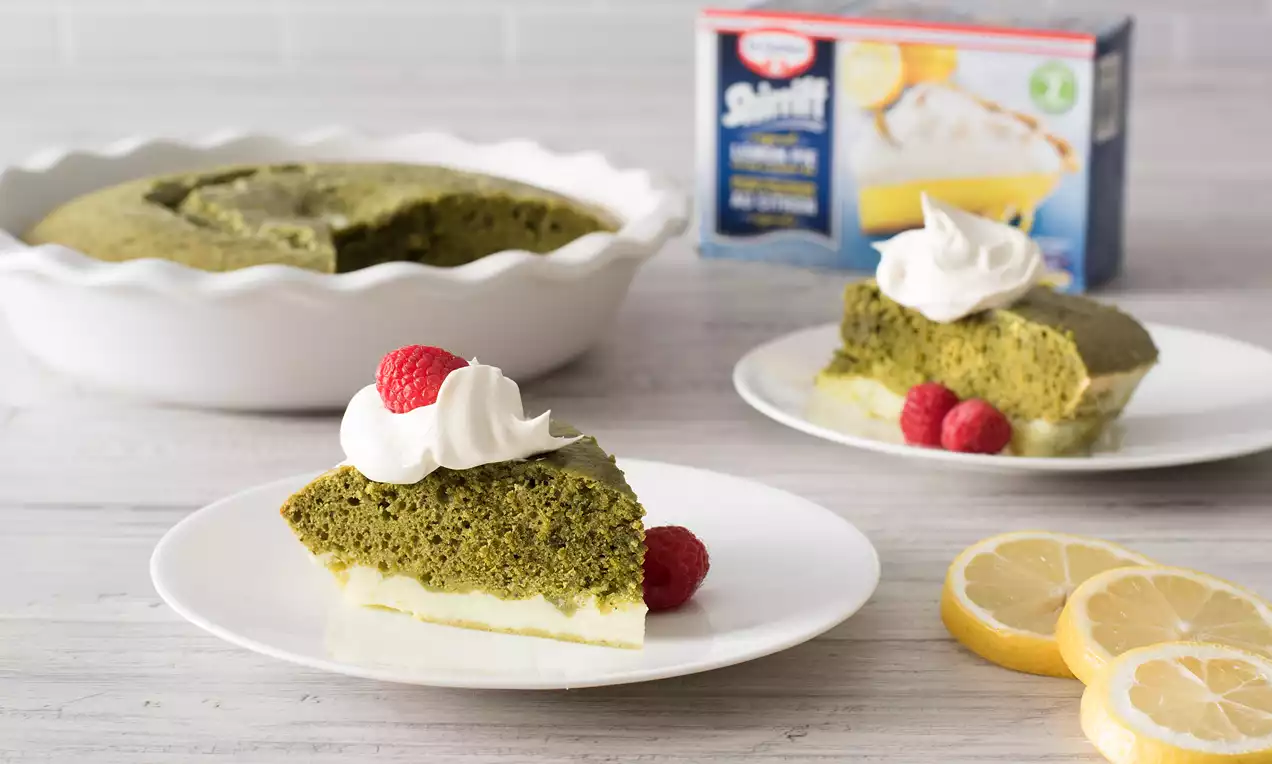 Matcha cake slice with vibrant green sponge, pale yellow pudding base, topped with white whipped cream and raspberries on a white plate