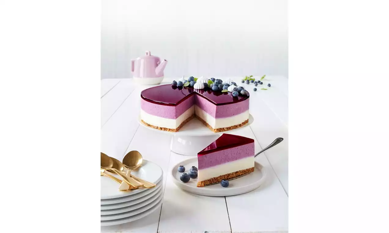 Blåbærkake med brun kjeksbunn, hvitt og lilla lag, mørkelilla gelétopp pyntet med blåbær og hvite blomster på hvitt bord med tallerkener