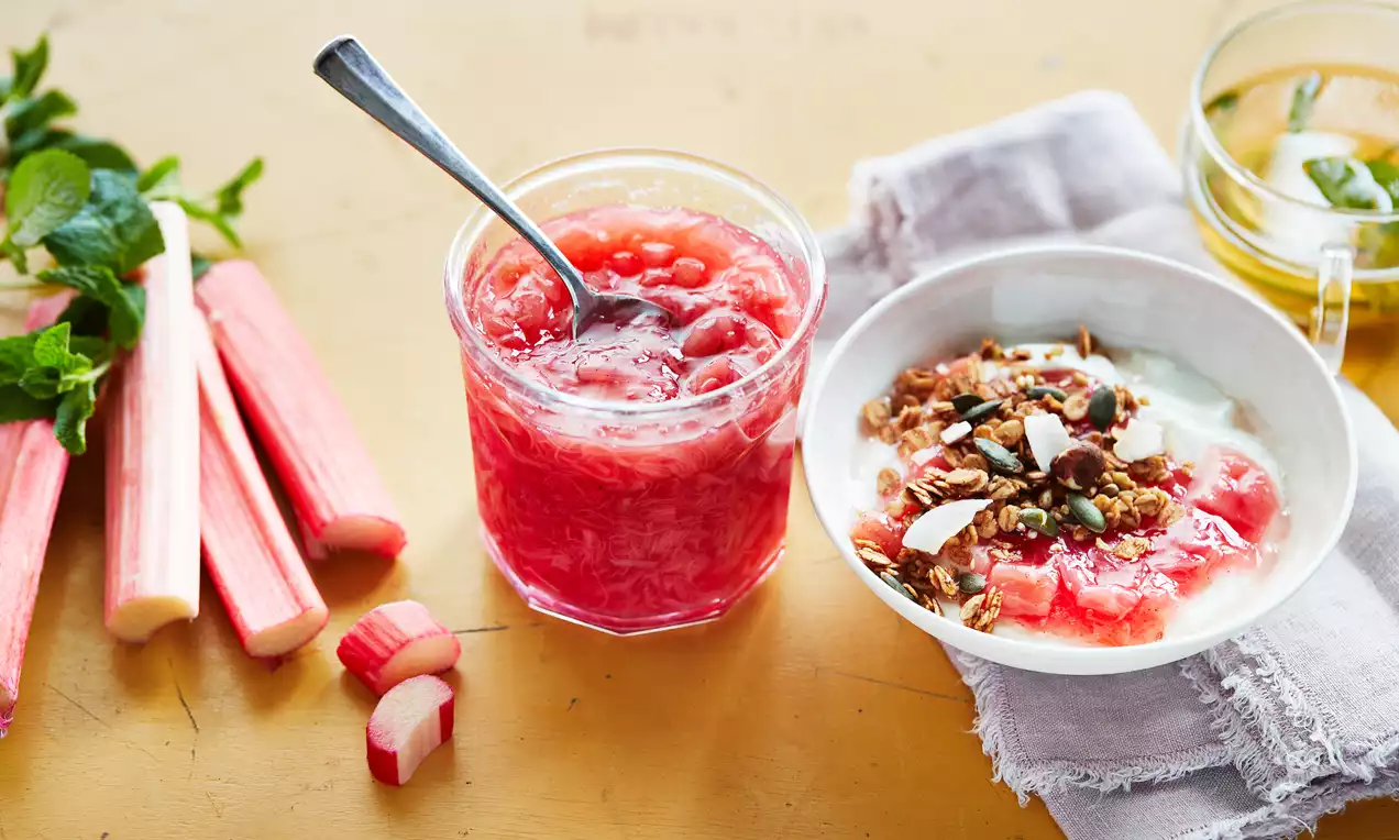 Vanille-rabarberconfituur in een glazen pot met lepel, felroze en glanzend, naast verse rabarberstengels en een schaaltje met yoghurt en granola