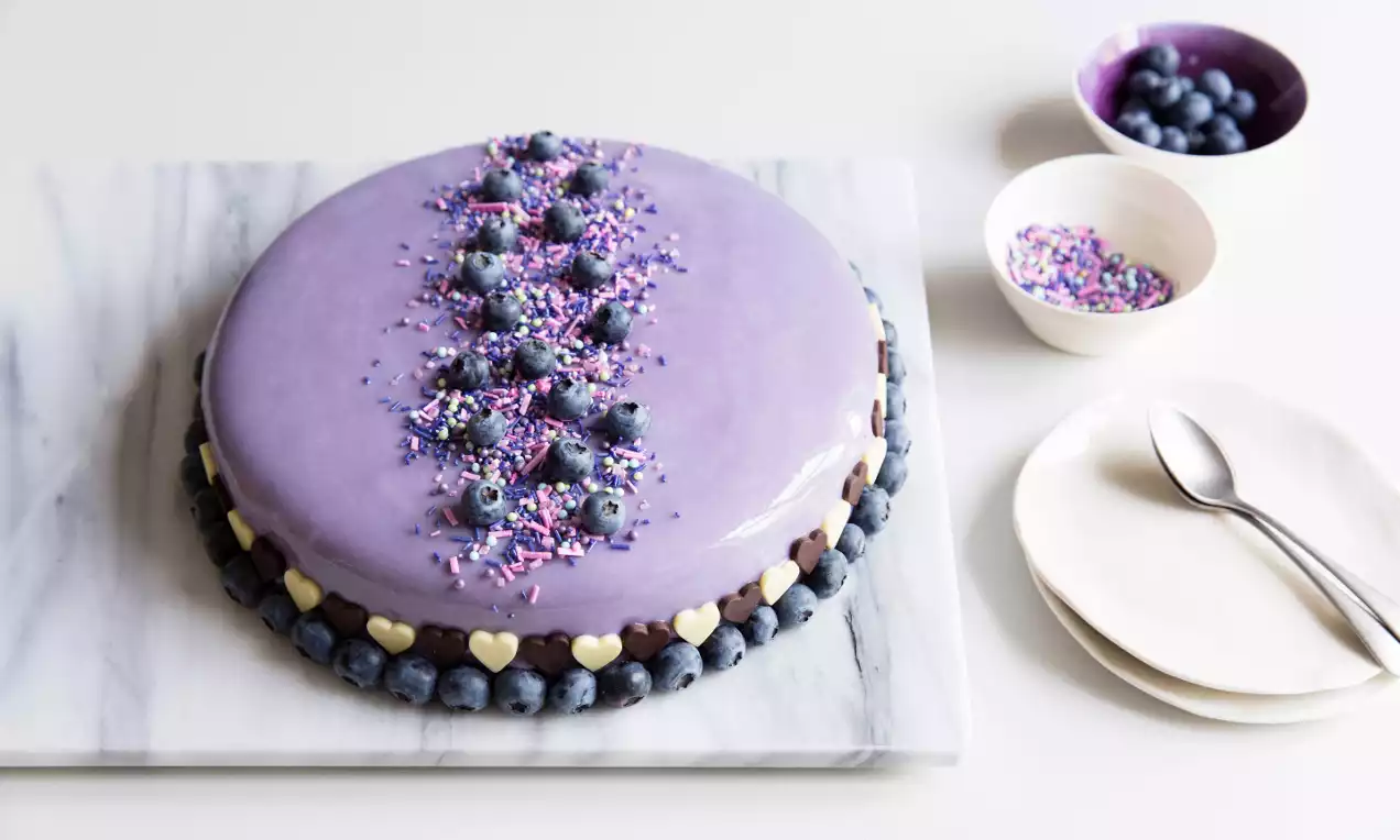 Lila fryst tårta med spegelblank yta, dekorerad med blåbär, strössel och vita hjärtan på en sockerkant, stående på en marmorskiva
