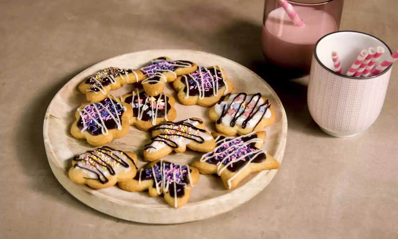 Bloemvormige zandkoekjes met goudbruine randen, kleurrijke glazuurstrepen en hagelslag op een licht houten bord, met roze melk en rietjes