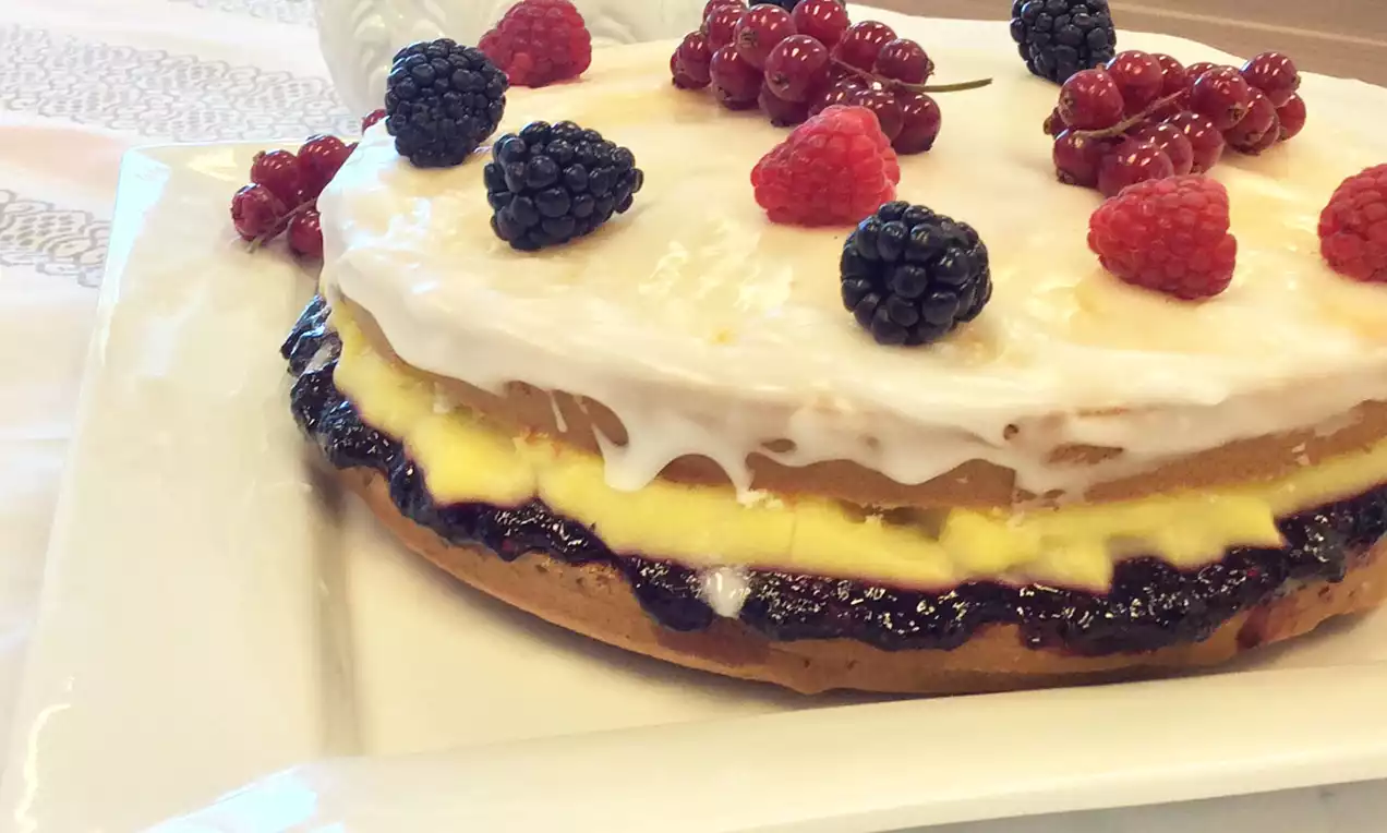 Gâteau à étages avec glaçage blanc, crème jaune et confiture sombre, garni de mûres, framboises et groseilles rouges sur une assiette blanche