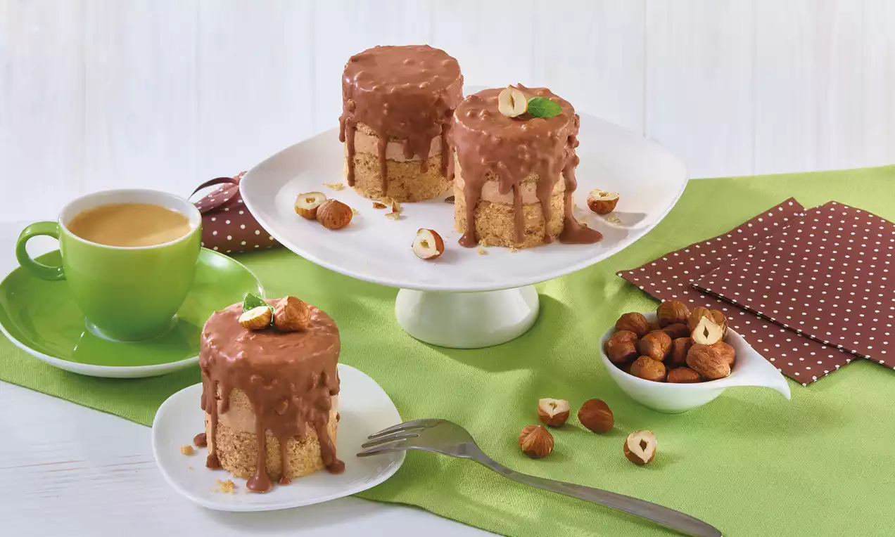 Nuss-Cremetörtchen mit beigem Boden und brauner Glasur, garniert mit Haselnussstücken auf weißem Teller, umgeben von grüner und brauner Deko