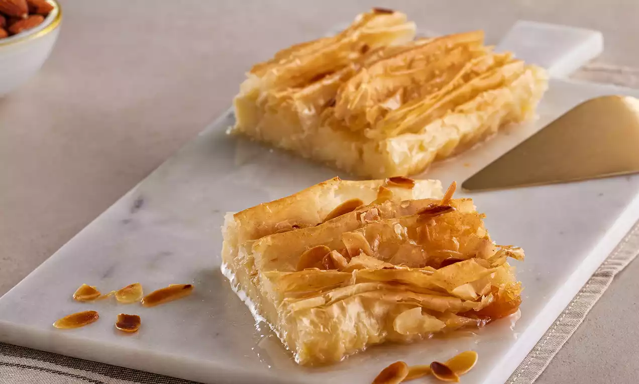 İnce kat kat yufkalar arasında kremalı keşkül dolgusu, üzeri kızarmış bademle süslü, şeffaf şurupla ıslatılmış, mermer tabakta sunulmuş