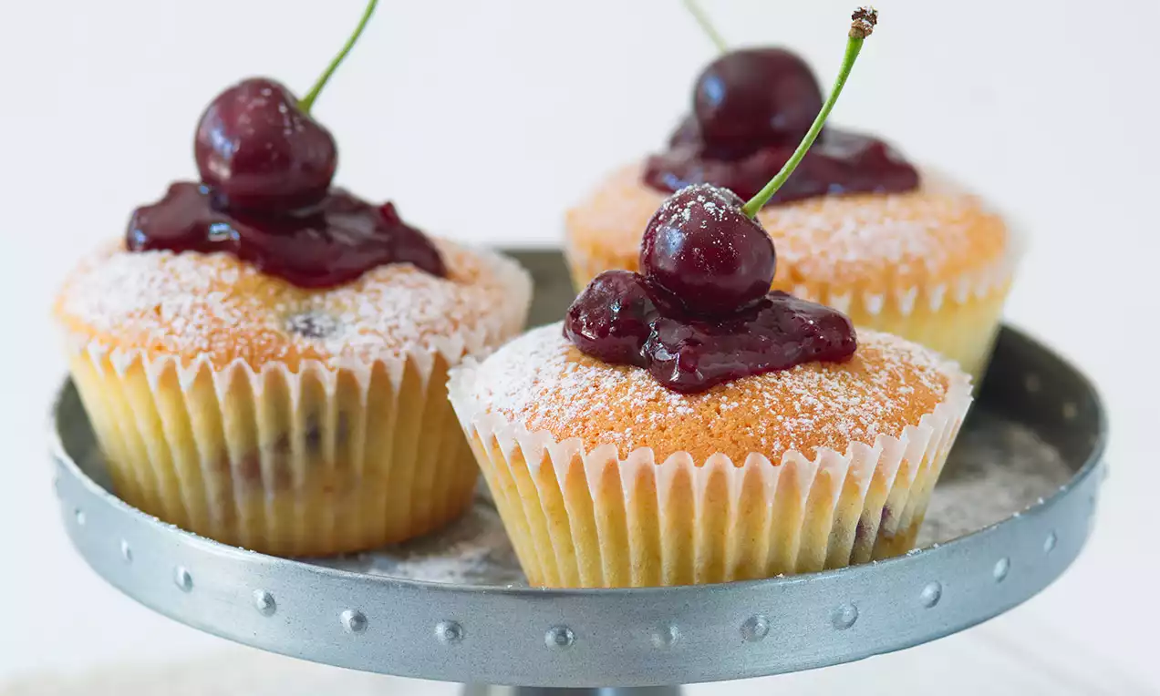 Višňové muffiny světle žluté barvy s jemnou nadýchanou texturou, posypané moučkovým cukrem, zdobené višňovým krémem a čerstvými višněmi na kovovém podnosu