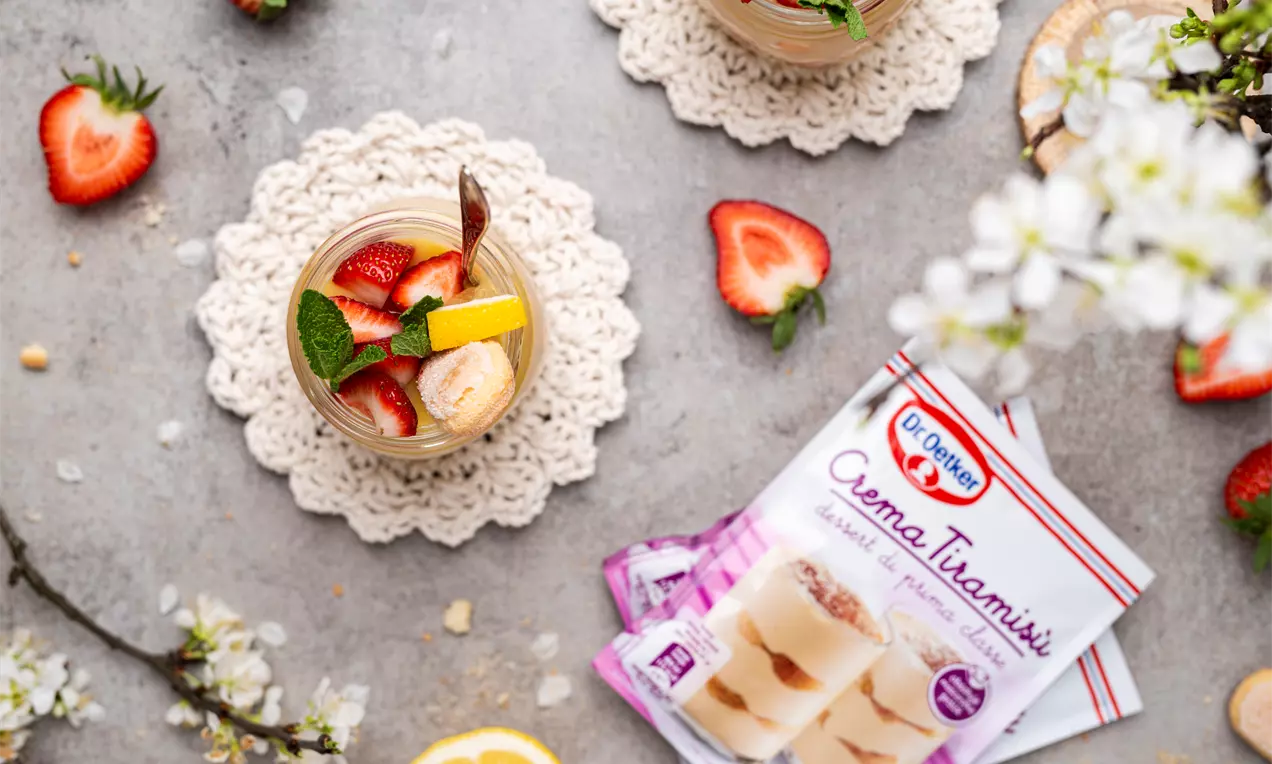 Stikliniame indelyje sluoksniuotas šviesiai kreminis tiramisu, papuoštas ryškiomis šviežiomis braškėmis, citrinos skiltele ir mėtos lapeliu