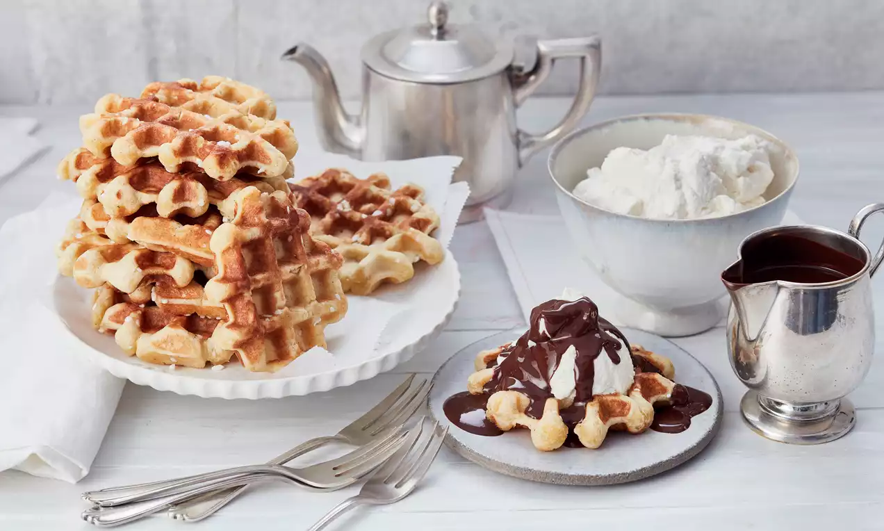 Belgische Waffeln goldbraun und weich, gestapelt auf einem weißen Teller, daneben Sahne in einer Schale und Schokoladensoße in einem Kännchen