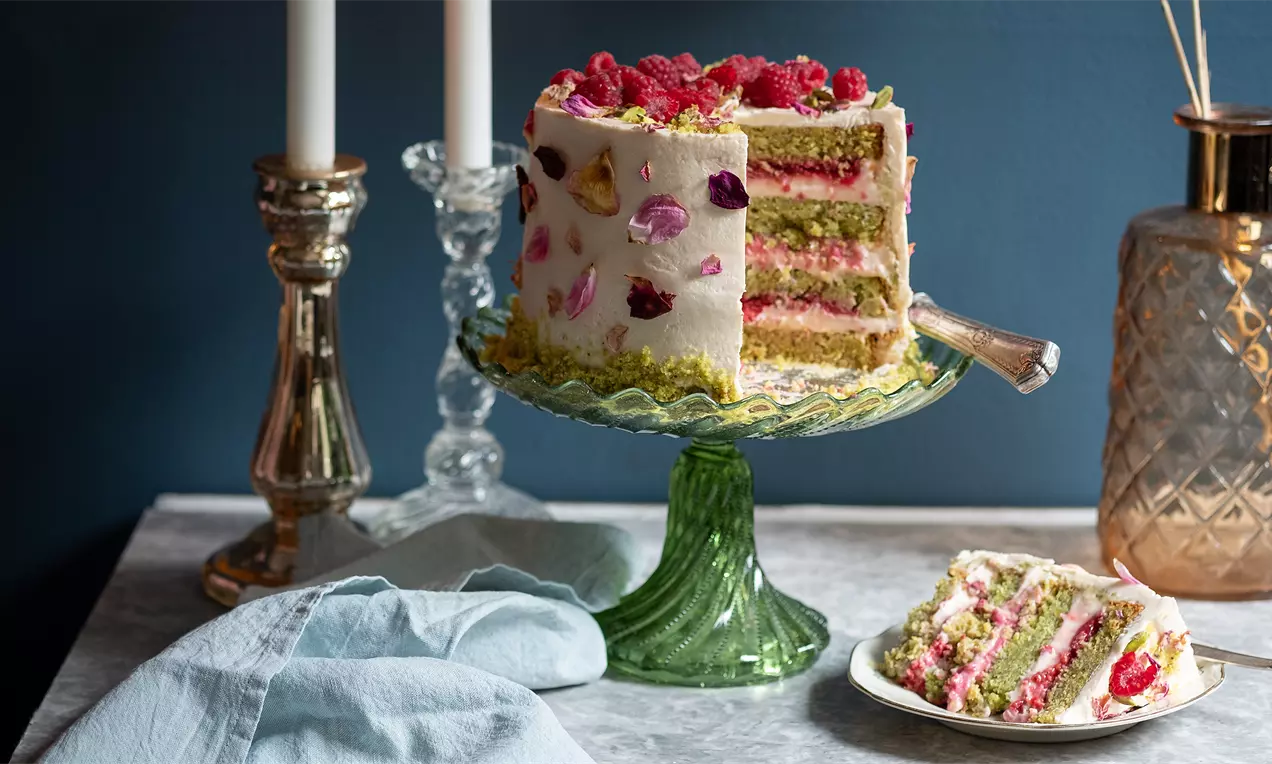 Висока торта с бели кремообразни страни, зелени и червени пластове, украсена с цели малини, шамфъстък и захаросани розови листенца