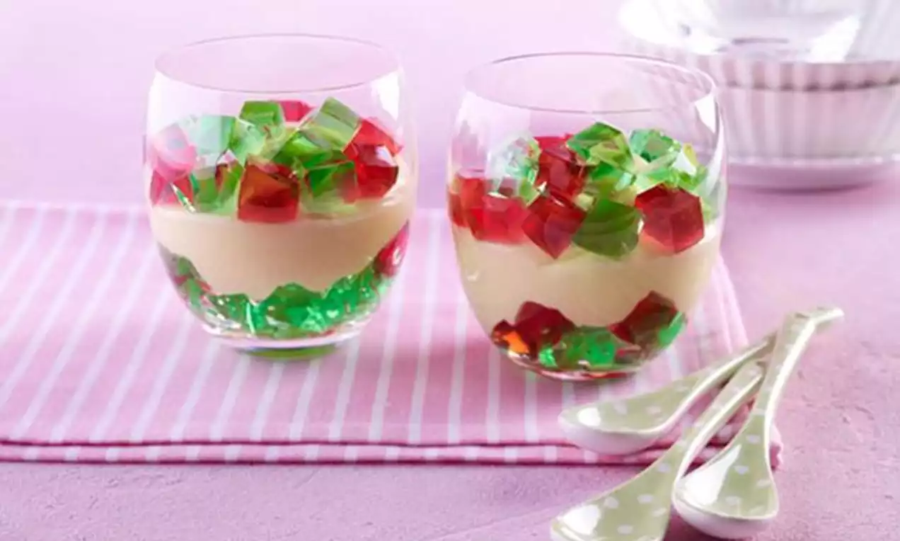 Zwei klare Gläser mit samtig gelbem Pudding und leuchtenden rot-grünen Geleewürfeln, auf rosa Streifenstoff mit gepunkteten Löffeln drapiert