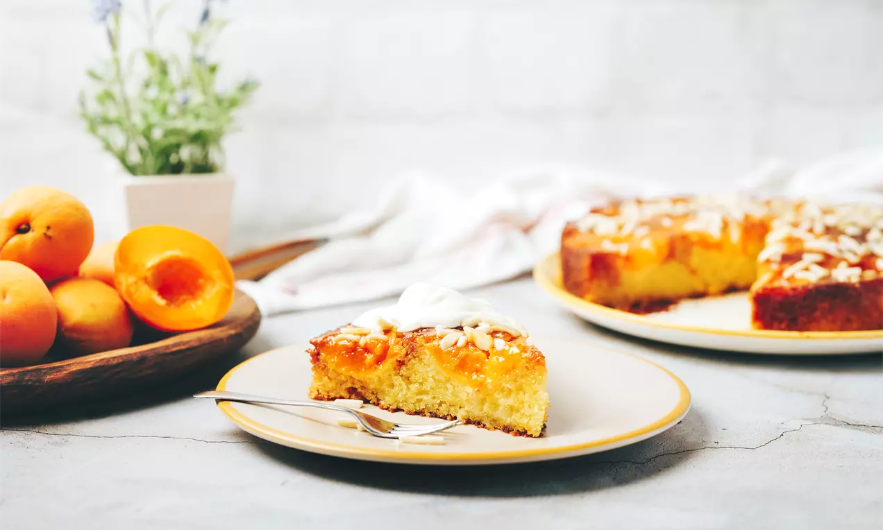 Златисто-жълт кейк с пухкава текстура, сочни резени кайсии и хрупкави филирани бадеми, сервиран върху порцеланова чиния