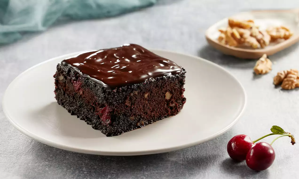 Yoğun çikolatalı dokusu ve parlak sosuyla kaplanmış, içerisinde ceviz parçaları ve vişne bulunan, beyaz tabakta sunulan kare dilim