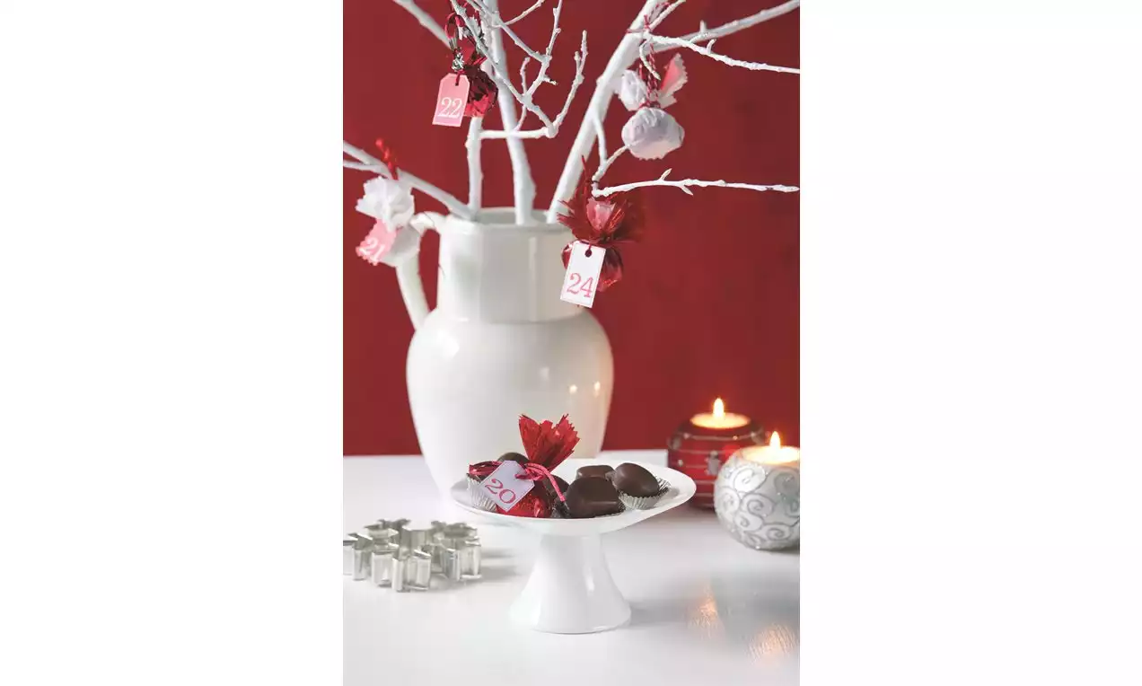 Pralinky tmavě hnědé barvy s lesklým povrchem na bílém stojanu, vedle červené dekorace, svíček a vázy s bílými větvemi