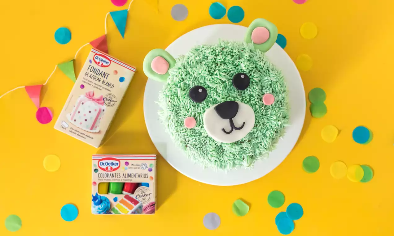 Tarta circular decorada como un oso verde con textura de crema, orejas rosas y hocico blanco sobre un plato blanco, rodeada de confeti