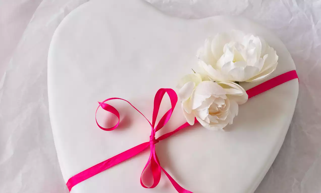Dort ve tvaru srdce s hladkou bílou polevou, zdobený dvěma krémově bílými květy a světle růžovou stuhou na světlejším podkladu