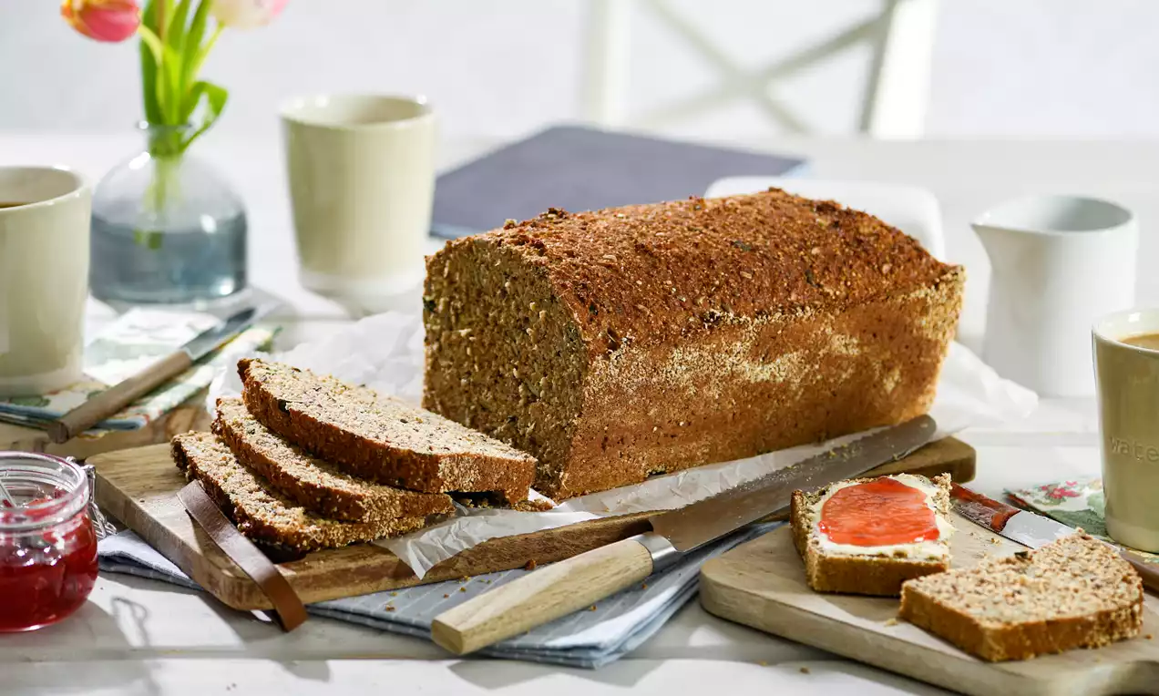 Ein länglicher Laib goldbraunes Low-Carb-Brot mit grober Textur, teils aufgeschnitten, dekoriert mit Aufstrichen auf einem hellen Holztisch