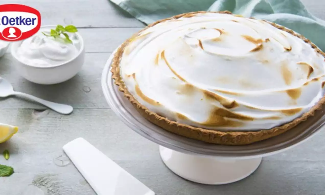 Tarte au citron avec une base dorée, garnie d'une meringue légère et dorée par endroits, sur un plat blanc près de couverts et citron