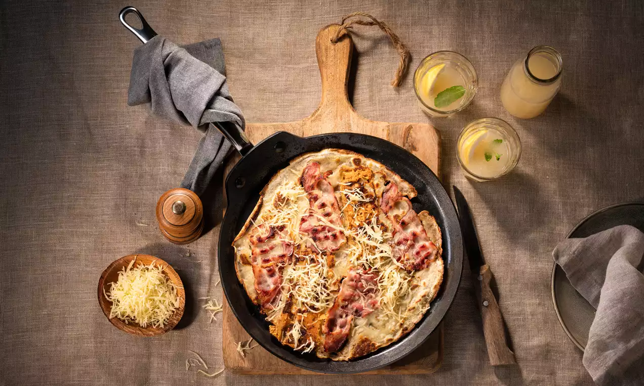 Spek en kaas pannenkoek goudbruin gebakken in een zwarte pan, bedekt met gesmolten kaas en knapperige spekreepjes op een houten plank