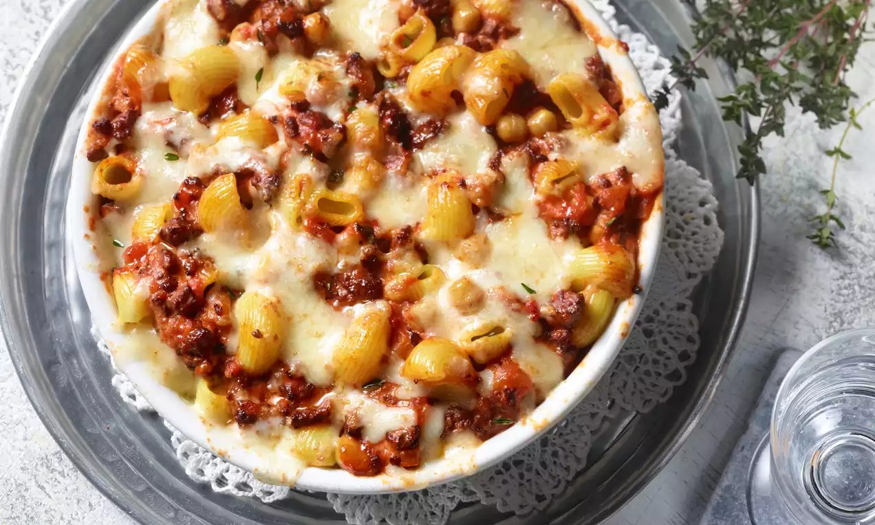 Ein goldbrauner Auflauf mit Röhrennudeln, Kichererbsen und geschmolzenem Käse in einer weißen Schale auf einem Spitzenuntersetzer