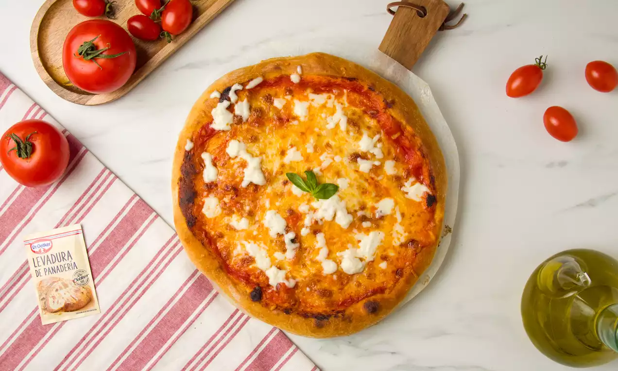 Pizza redonda con costra dorada, base de tomate rojo intenso, queso fundido blanco y albahaca en una mesa con tomates y aceite