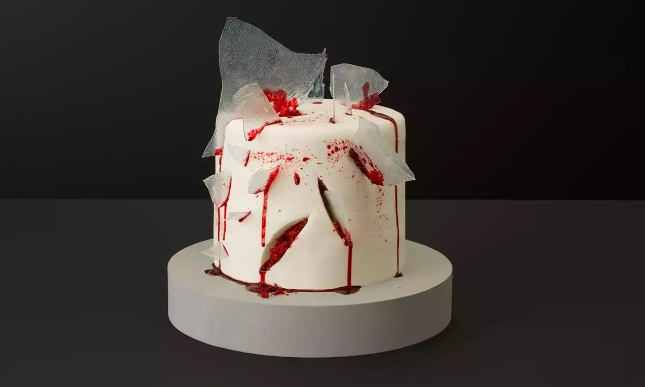 Tall white cake with smooth icing, jagged transparent shards, and dripping red glaze pooling at the base on a pale grey stand