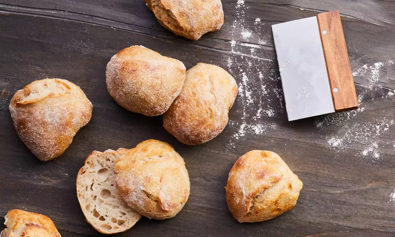 Gyldenbrune surdeigsboller med rustikk skorpe og luftig innside ligger spredt på et mørkt trebord ved siden av en deigskrape med treskaft