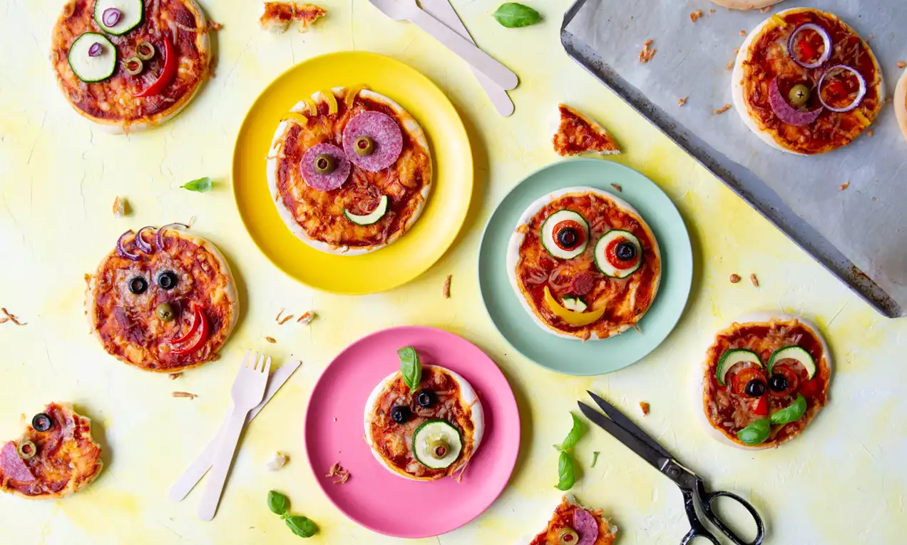 Kleine ronde pizza's met kleurrijke topping in decoratieve gezichten, gerangschikt op gekleurde borden naast basilicum en keukengerei
