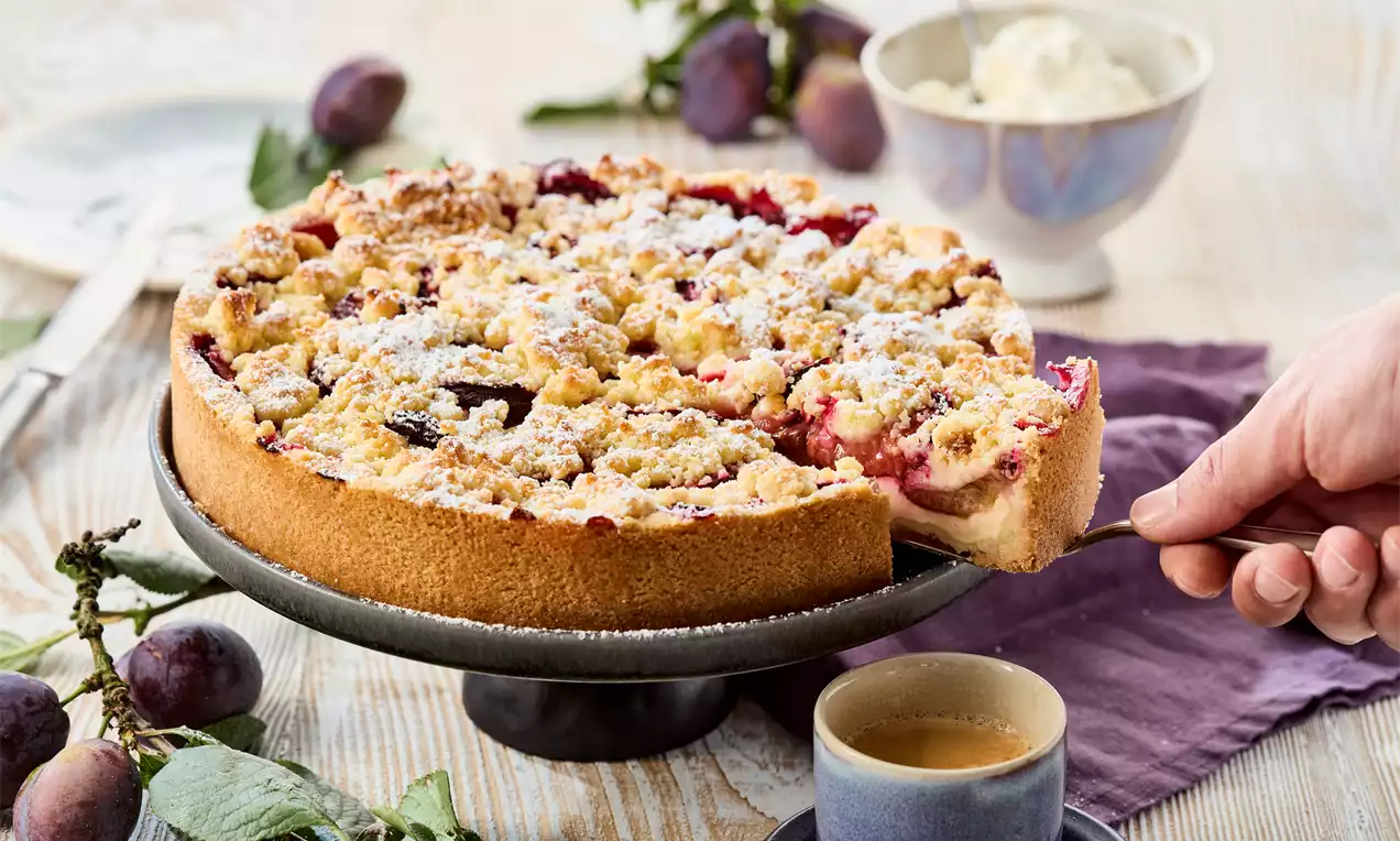Pflaumen-Pudding-Kuchen mit hellbraunem Rand, goldenen Streuseln und roten Pflaumenstücken, bestäubt mit Puderzucker auf einer grauen Kuchenplatte