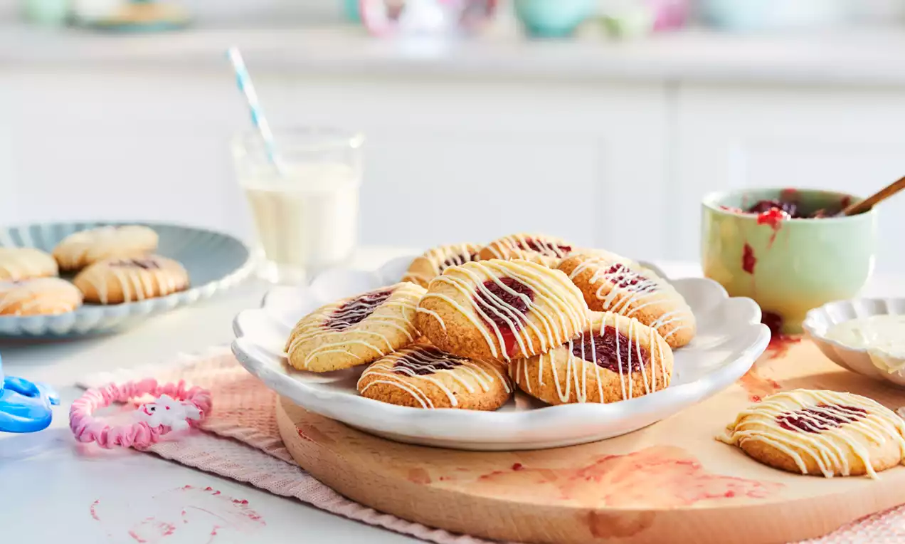 Golden thumbprint cookies with a glossy red jam center and white icing drizzle arranged on a scalloped white plate on a wooden board