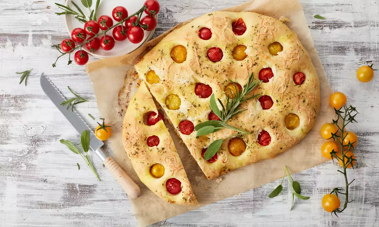 Goldbraunes, flaches Pizzabrot mit roten und gelben Kirschtomaten, angerichtet auf Backpapier mit frischen Kräutern und einem Messer