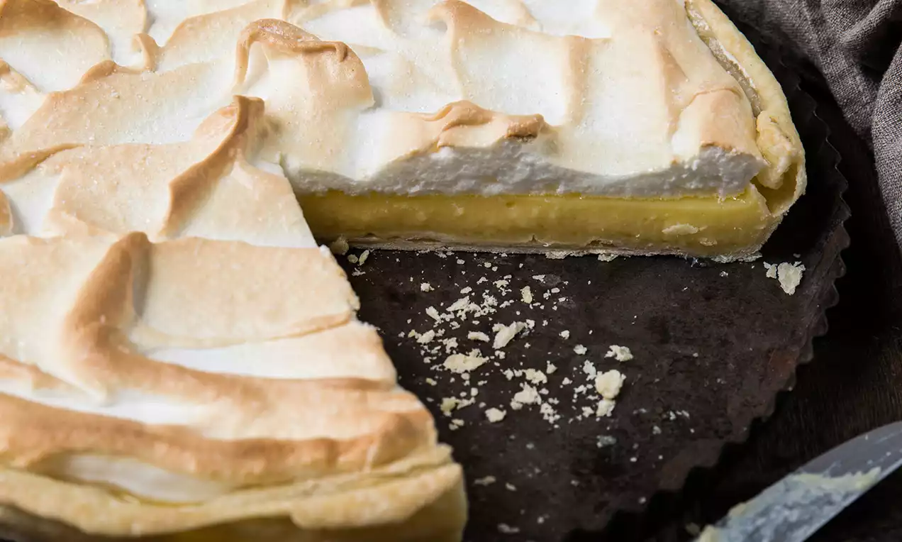 Lemon Meringue Pie mit hellgelber Zitronenfüllung, goldbrauner Baiserhaube und Mürbeteigkruste, angeschnitten in einer dunklen Tarteform