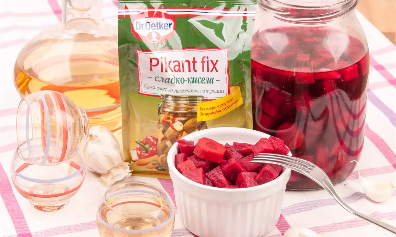 Червени парчета туршия с лъскава текстура, подредени в бяла купичка до голям буркан с наситен червен маринад върху раирана покривка