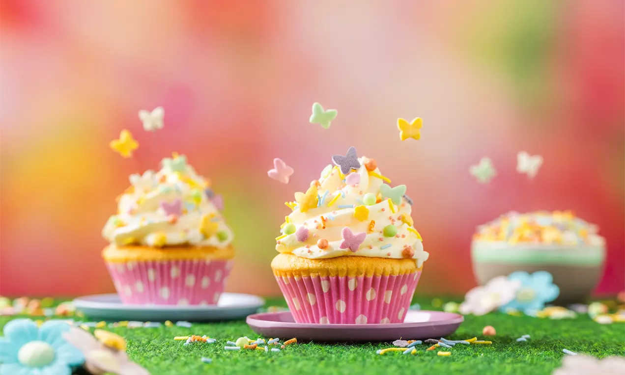 Cupcakes in roze vormpjes met luchtige witte topping, gekleurd met vlindervormige suikerstrooidecoratie op groen gras met bloemen