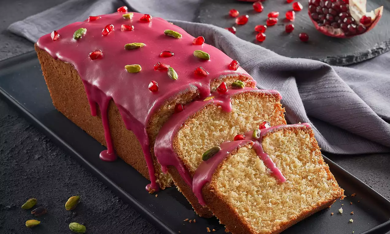 Narlı kek, parlak pembe nar sosu ve taneleriyle süslenmiş, ince dilimlenmiş, yanında antep fıstıkları serpilmiş siyah tabakta