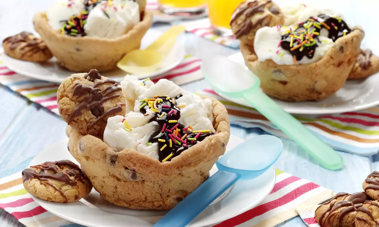 Cookie bowls with a golden-brown baked surface holding scoops of white ice cream topped with chocolate sauce and colourful sprinkles on striped cloth