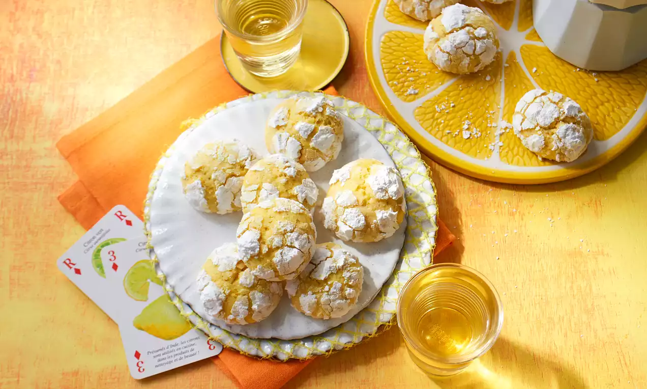 Zitronen-Crinkle-Cookies mit goldgelber, rissiger Oberfläche und weißem Puderzucker auf dekorativen Tellern neben Gläsern mit Getränk