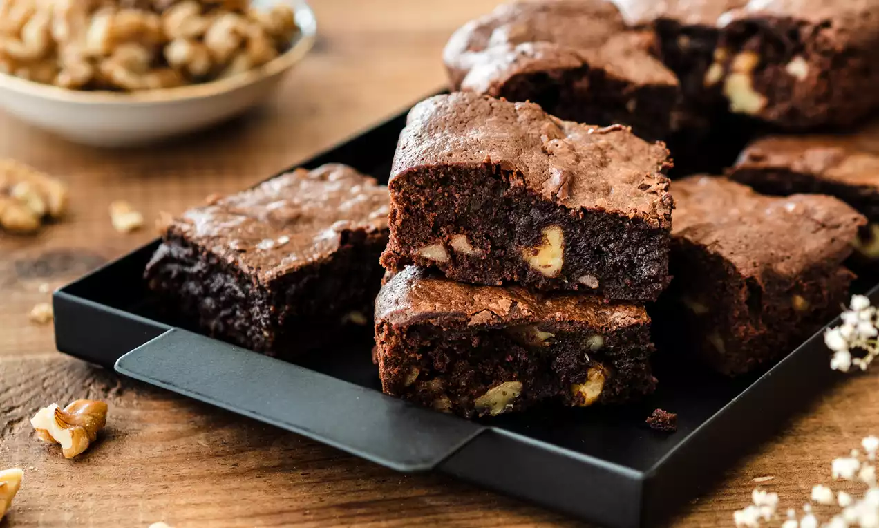 Kwadratowe kawałki intensywnie czekoladowego brownie z widocznymi kawałkami złocistych orzechów, ułożone na czarnej metalowej tacy