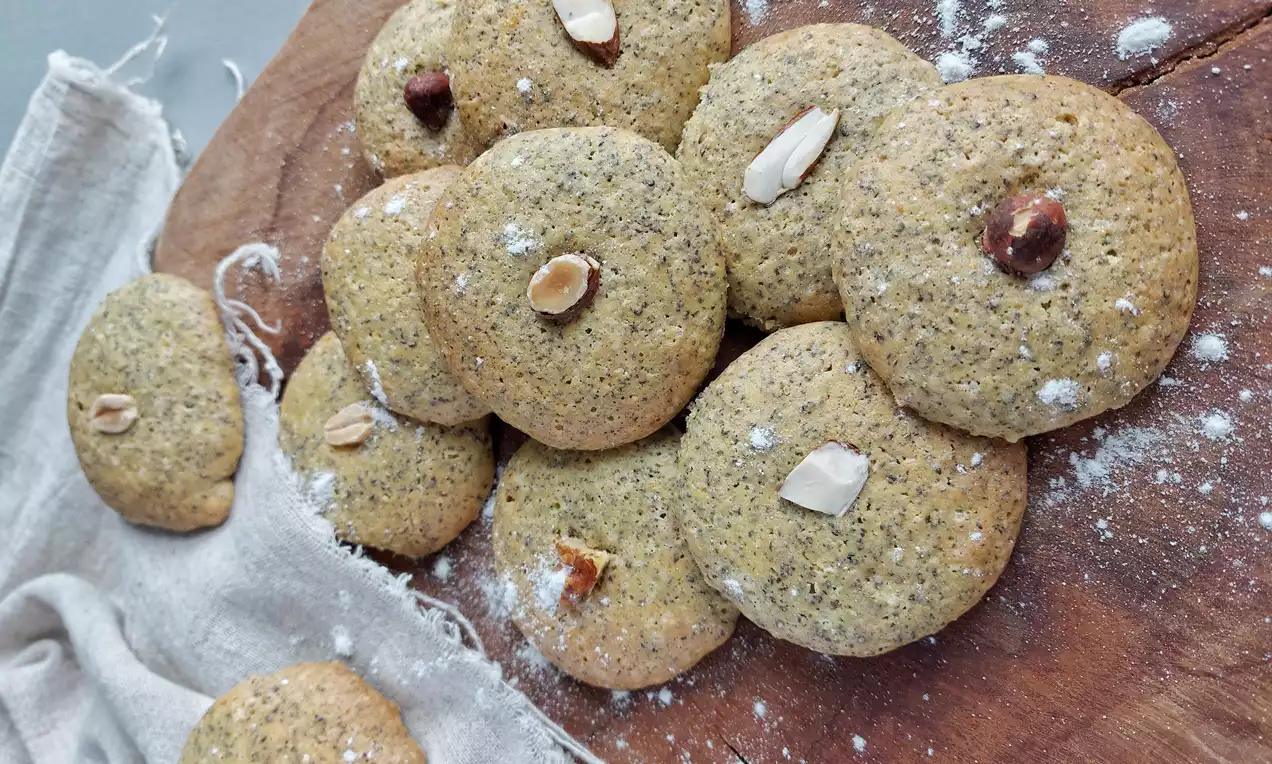 Kulatá světlá maková kolečka s jemnou zrnitou texturou, zdobená celými oříšky a plátky mandlí na dřevěném podkladu vedle látky